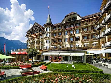 Grand hotel exterior with "Lauberhorn" sign, garden terrace, and mountains under a blue sky.