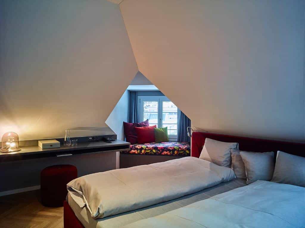 Bedroom in an attic space with a low bed, built-in desk, and window nook with colorful pillows.