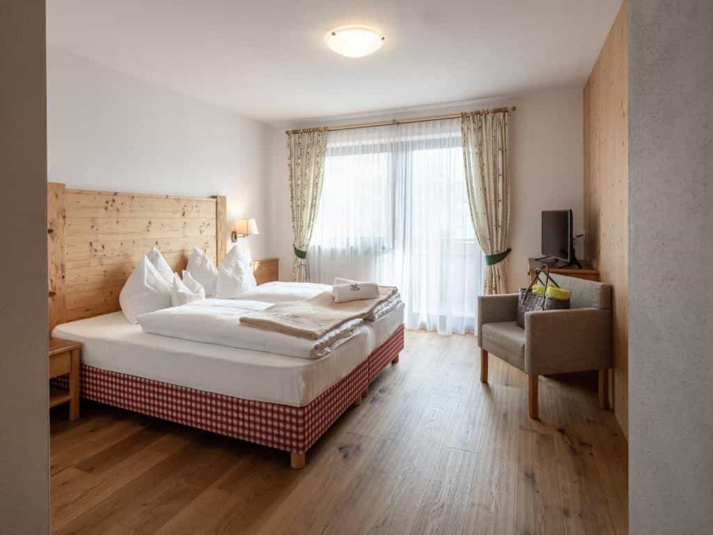 Hotel room with wooden headboard, white bedding, armchair, and sheer patterned curtains.