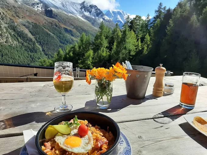 Outdoor dining table with food, drinks, and flowers overlooking alpine mountains and pine forest