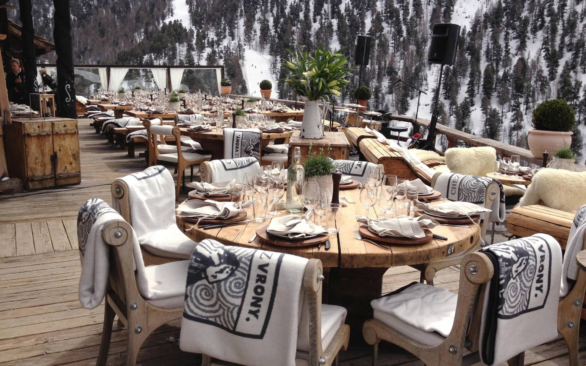 Outdoor restaurant dining terrace with wooden tables and chairs overlooking a snowy alpine forest slope