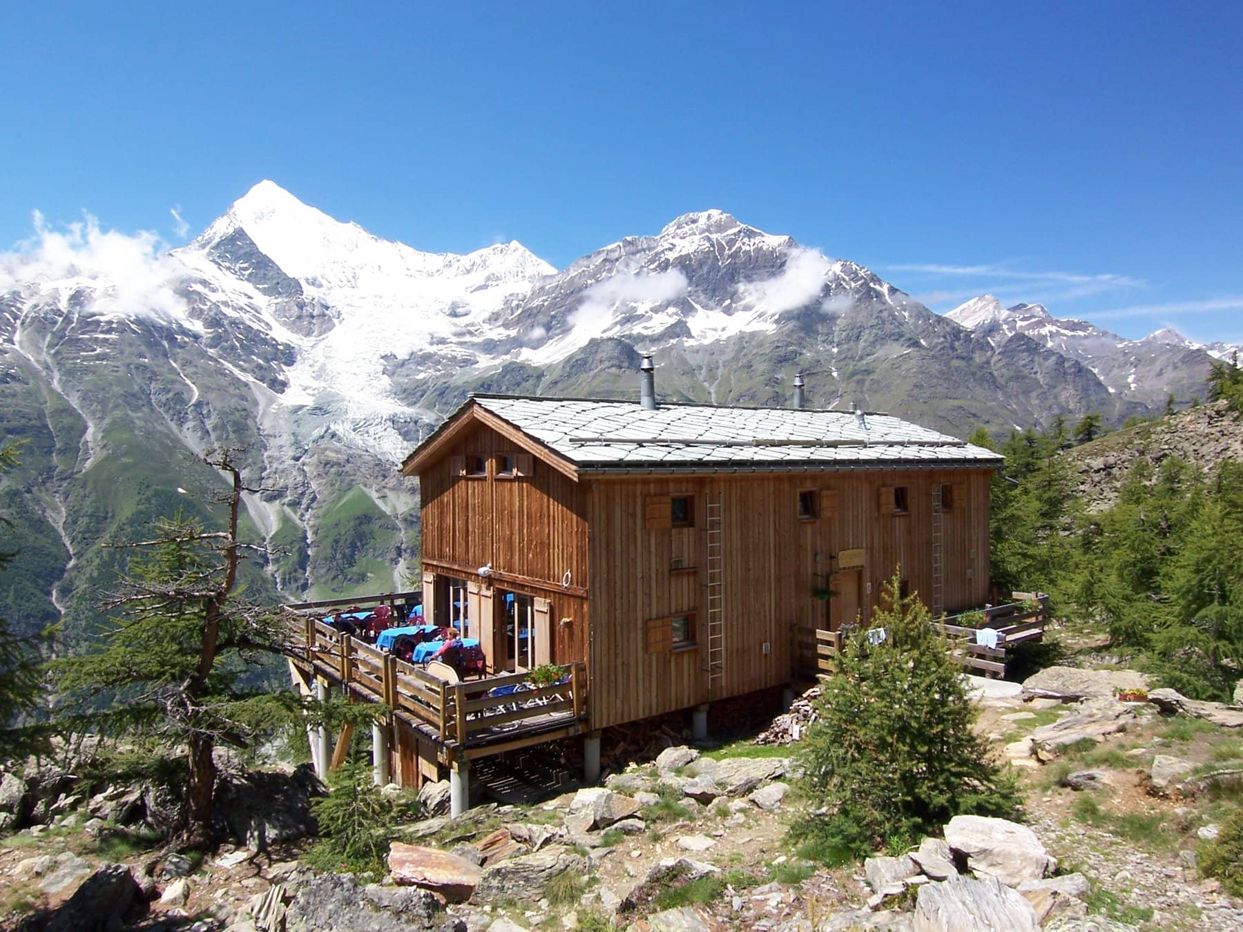 Wooden alpine hut with outdoor seating terrace against snowy mountain peaks