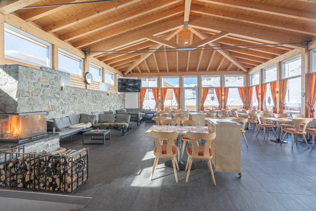 Restaurant interior with wooden ceiling, stone fireplace, and large windows overlooking snow