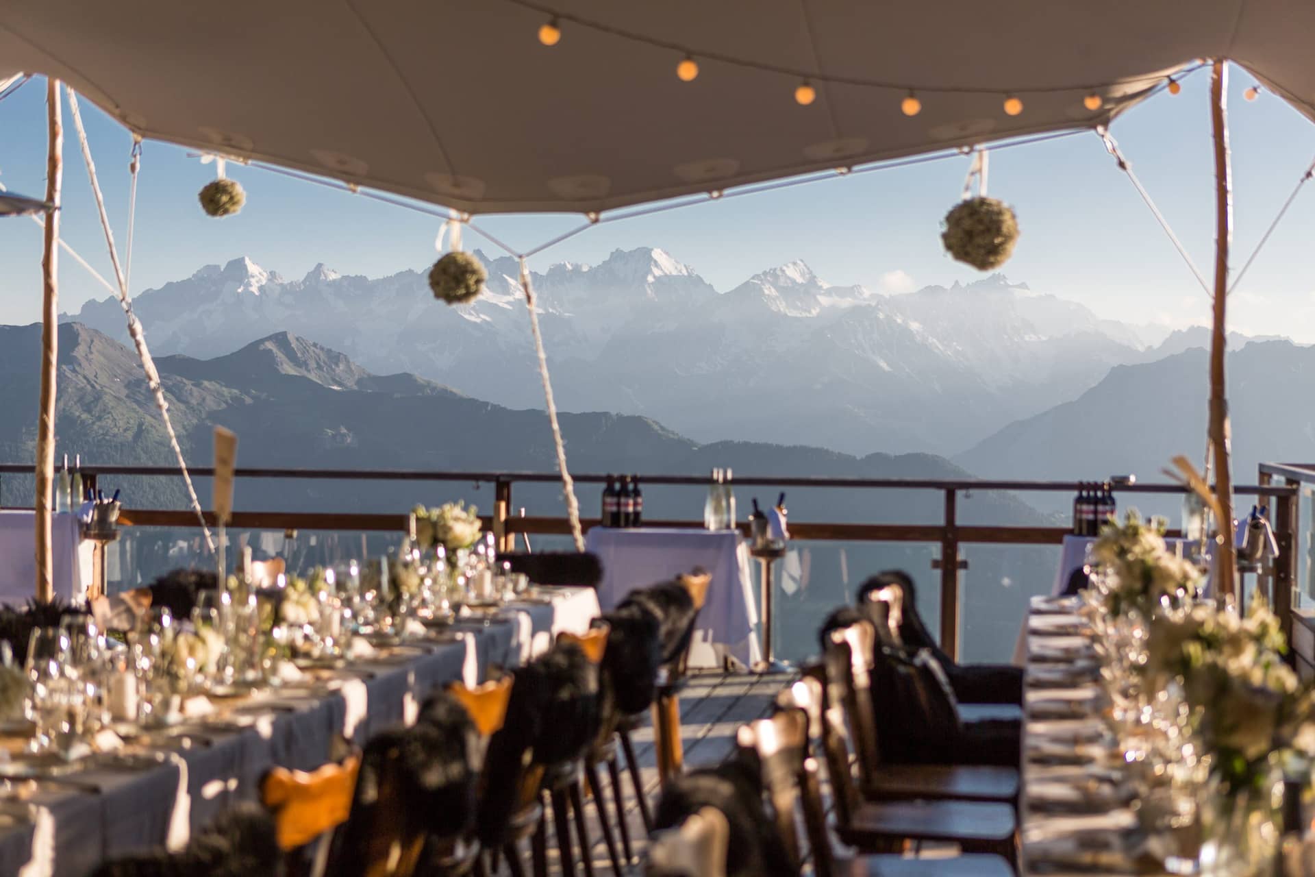Outdoor dining tables set with glasses and floral arrangements overlooking snow-capped alpine mountains under a tent canopy