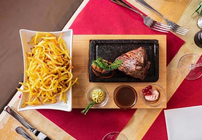 Grilled steak served on stone plate with french fries, sauce, and side dishes on wooden board