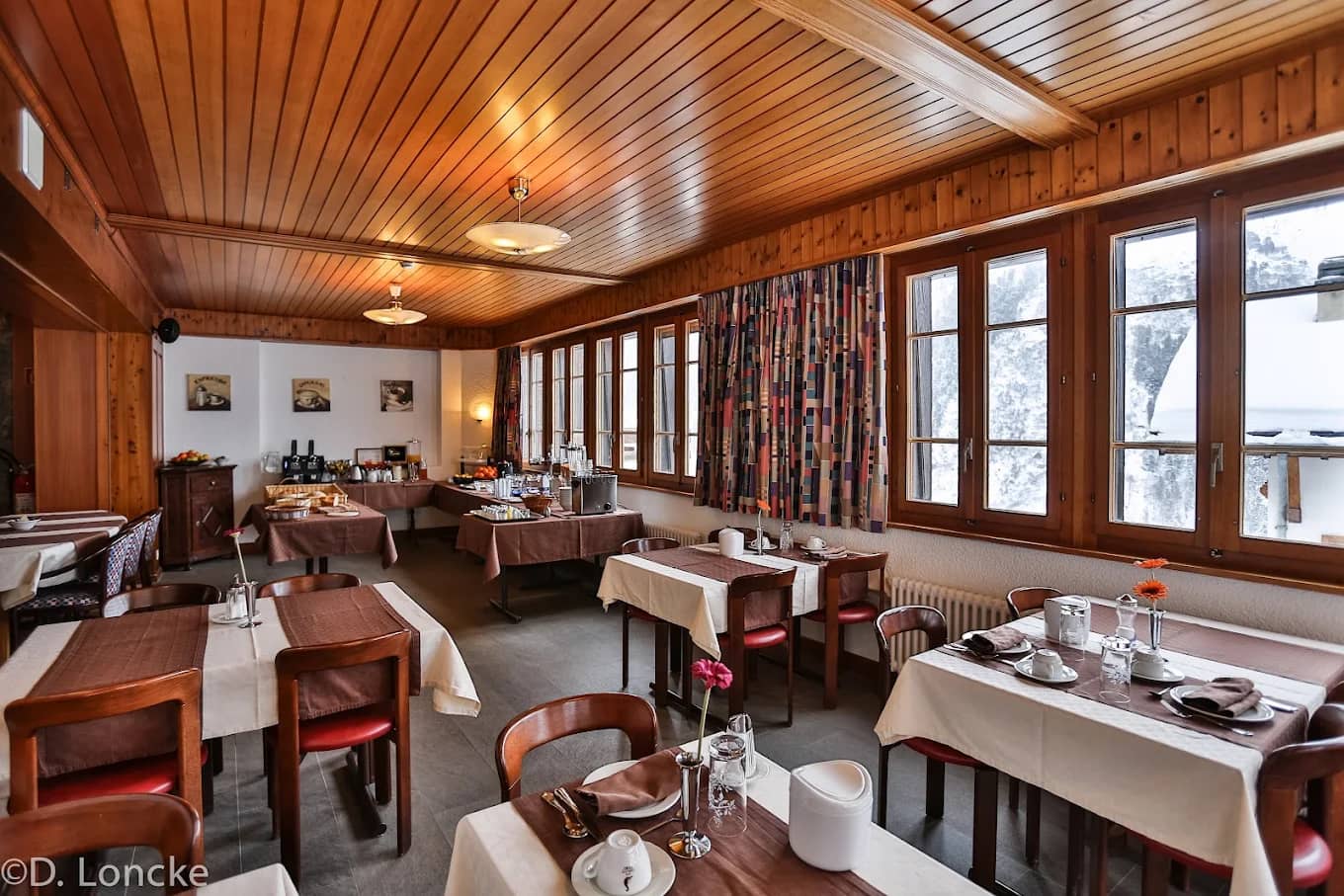 Hotel dining room with wooden panel ceiling, tables set with white tablecloths, and large windows overlooking snow