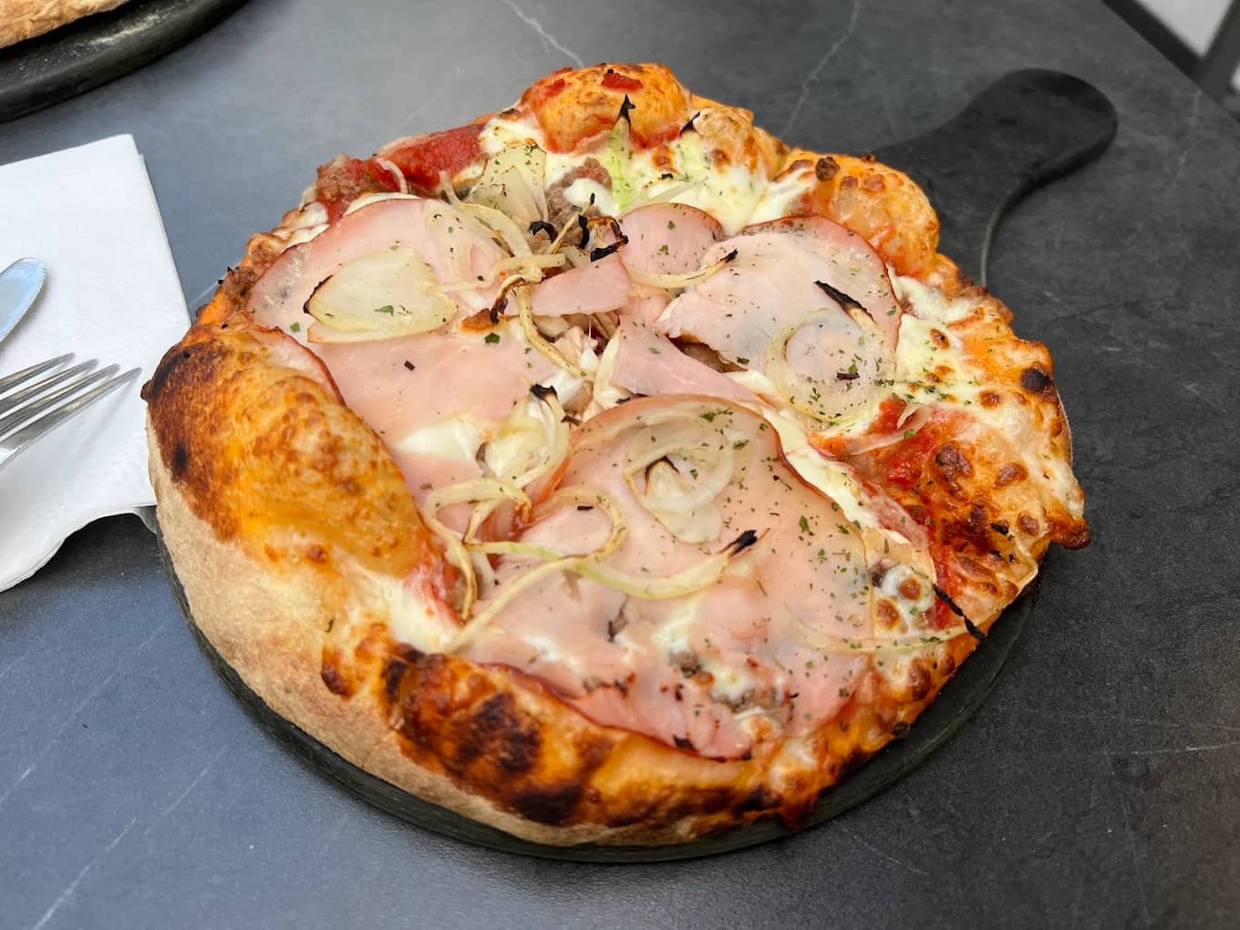 Wood-fired pizza topped with ham, cheese, and onions on a black serving paddle