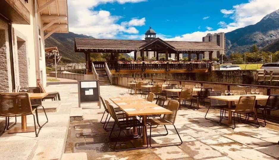 Outdoor cafe terrace with wooden tables and chairs facing a mountain stream and bridge