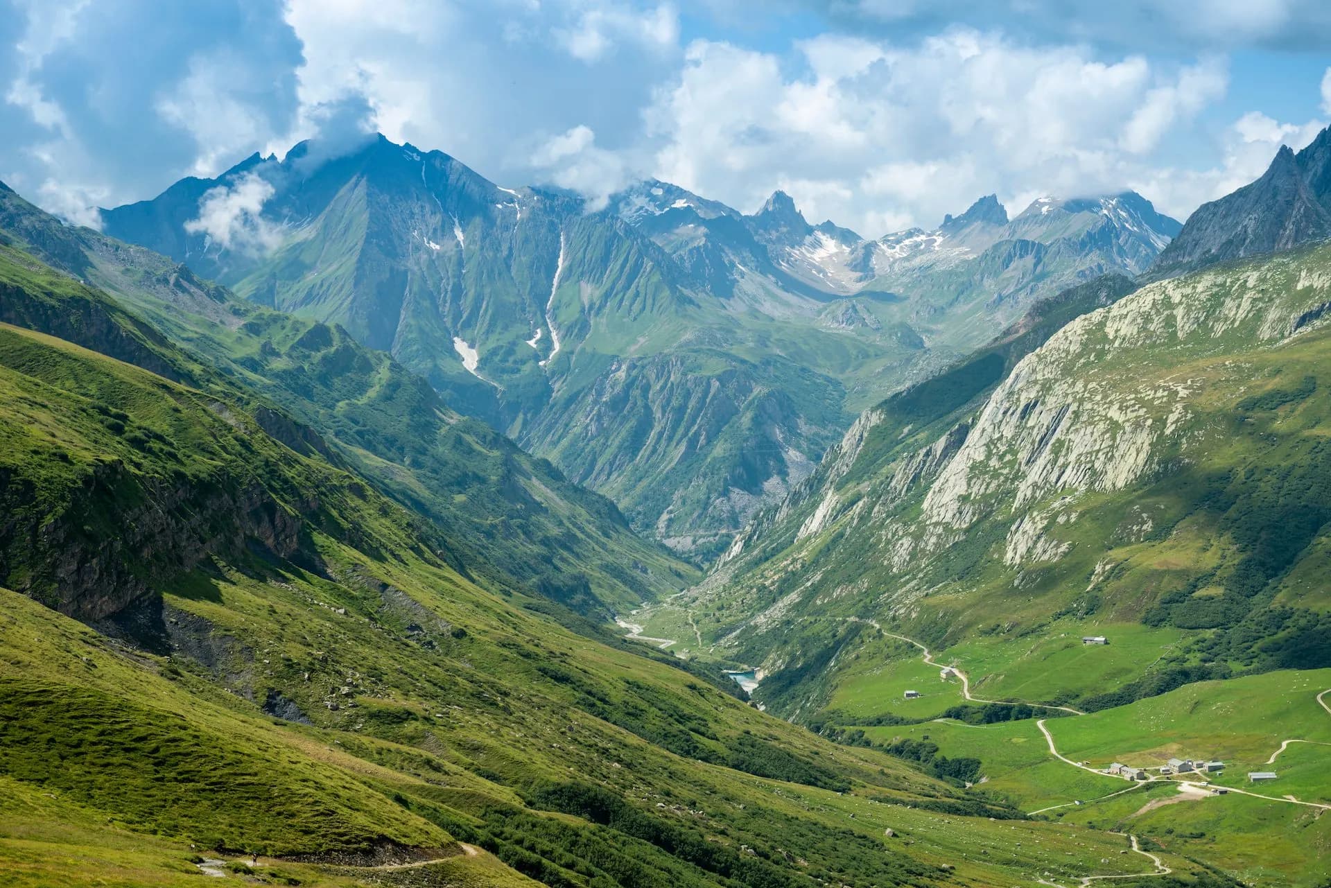 Green alpine valley with steep slopes, rugged mountains, and scattered buildings under a cloudy blue sky.