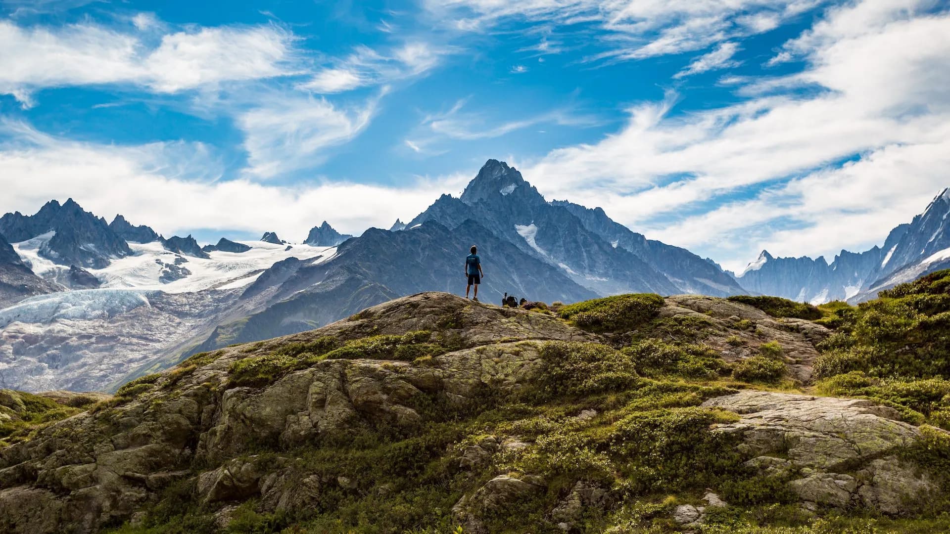 Hiker standing on rocky outcrop overlooking massive snow-capped mountains under blue sky