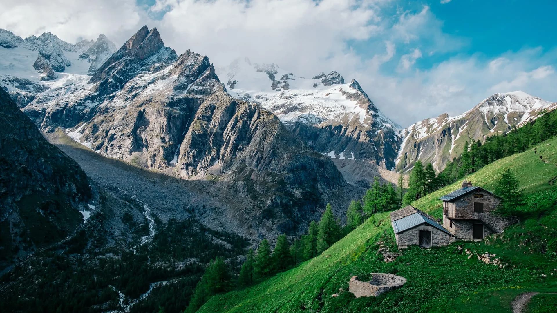 Stone refuge huts on a steep green slope below snowy alpine mountains, Refuge Bonatti.