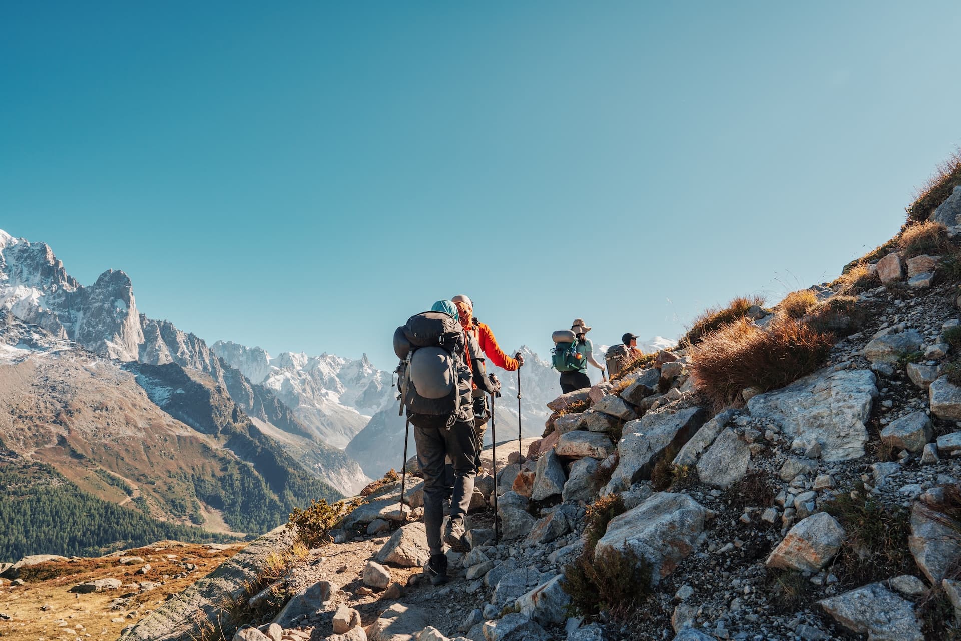 Hikers with large backpacks ascend a rocky alpine trail toward snow-capped mountains under a clear blue sky.