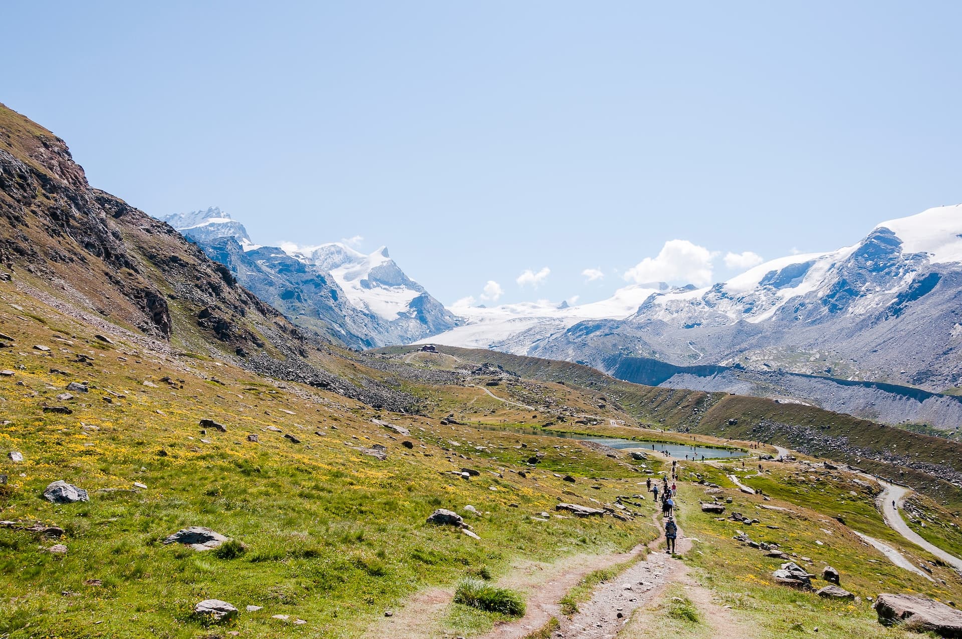 Hikers on trail toward Stellisee with massive snow-capped mountains and glacier backdrop