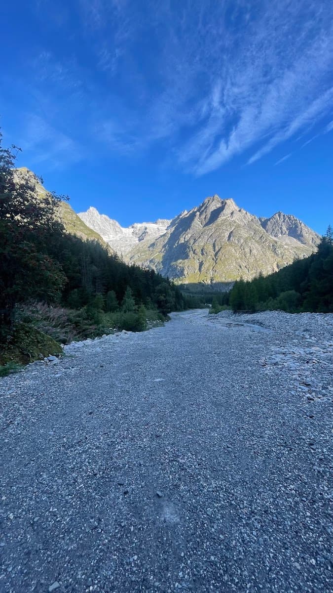 Wide, dry riverbed of pebbles leading toward towering, partially snow-capped mountains under a blue sky.