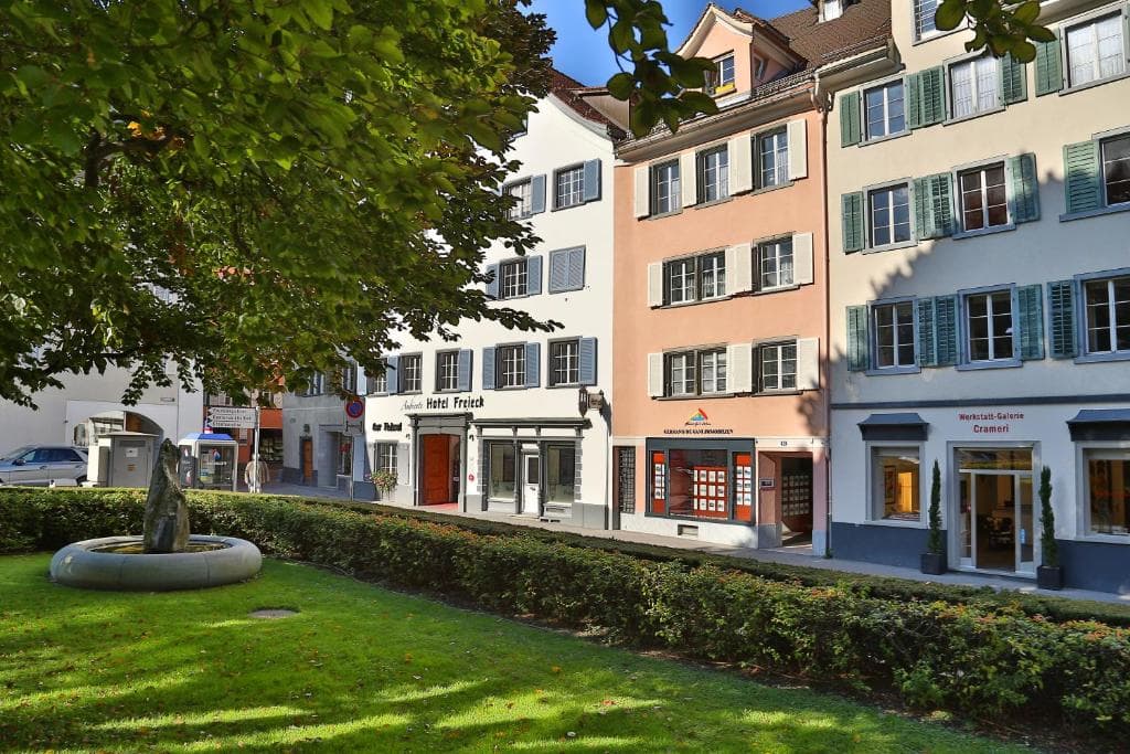 Hotel Freieck facade next to shops, viewed across a sunny lawn with a stone fountain.