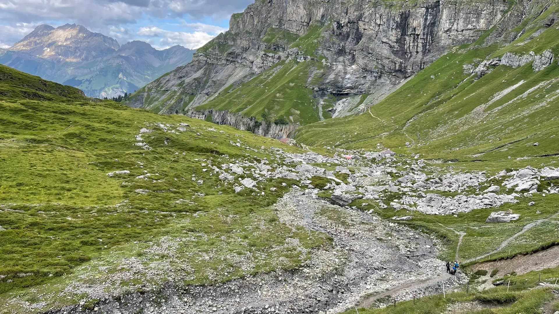 Hikers on a trail in a green alpine valley with rocky slopes and distant mountain peaks.