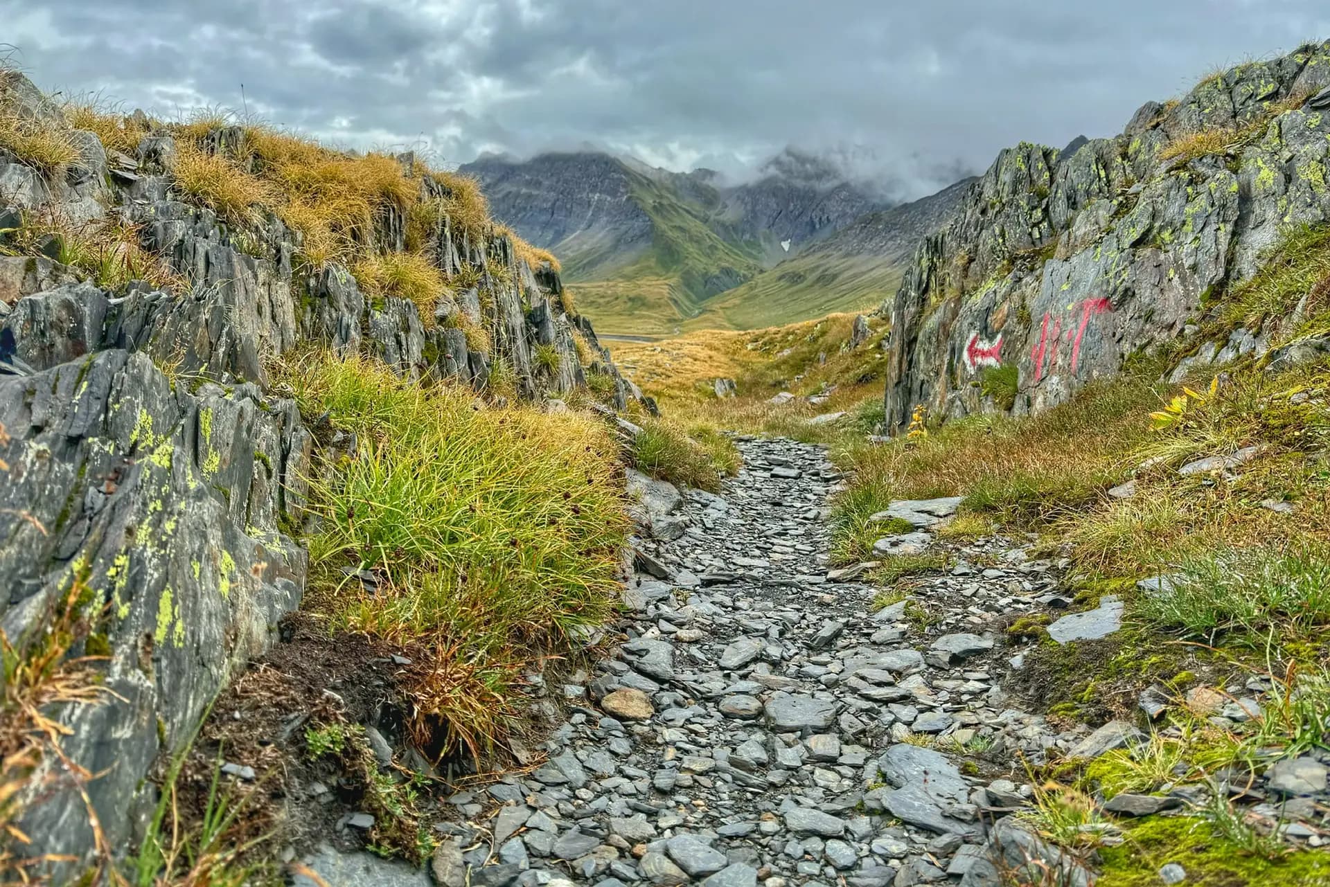 Rocky hiking trail leading through a mountain pass with alpine meadows and cloudy peaks.