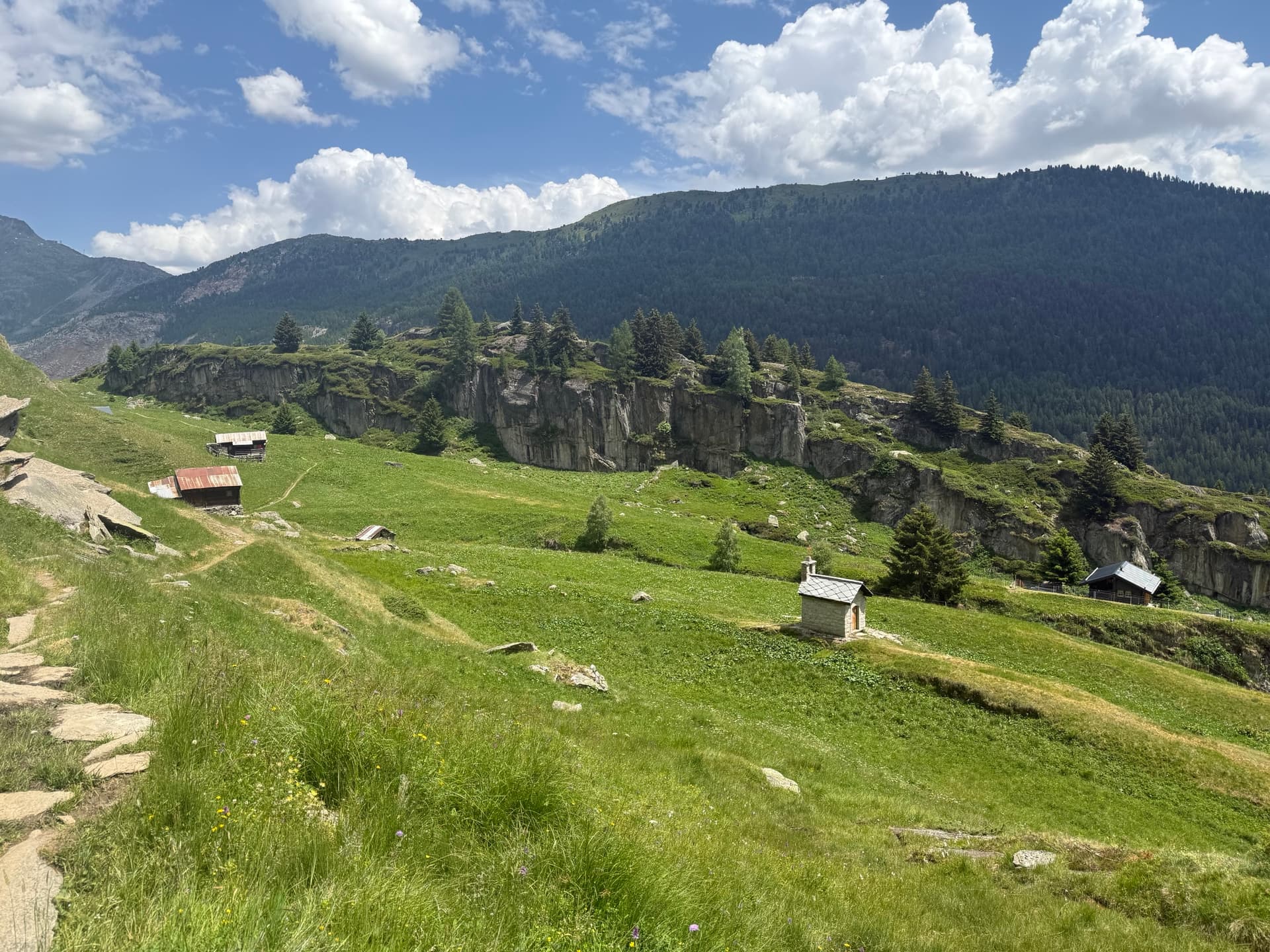 Small stone chapel and wooden huts on green alpine pasture with rocky cliffs and mountains