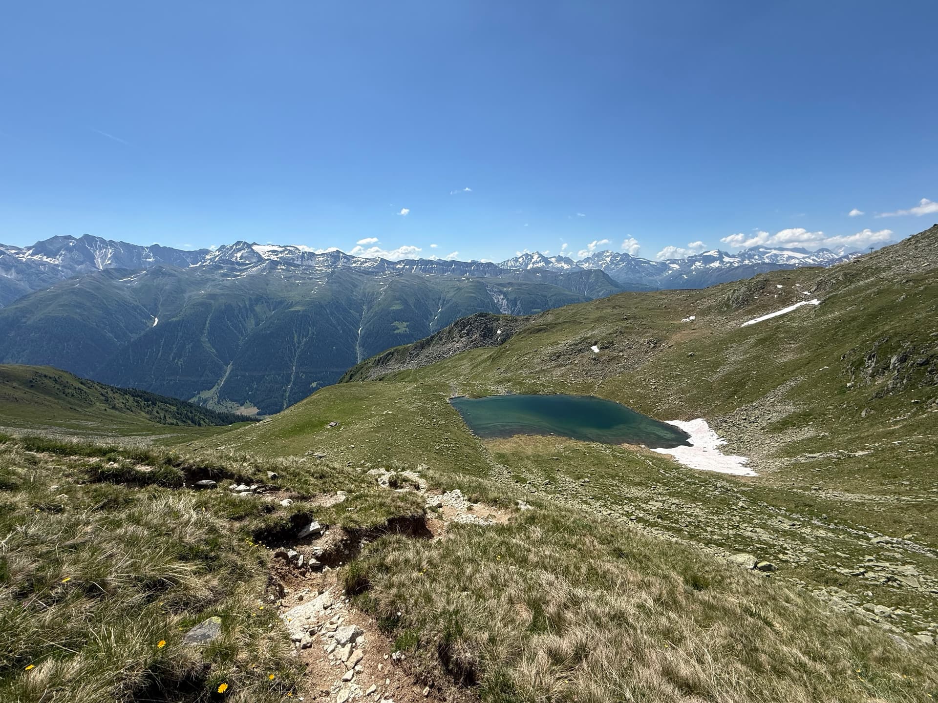 Trail leading to a small mountain lake with distant snow-capped peaks under a blue sky