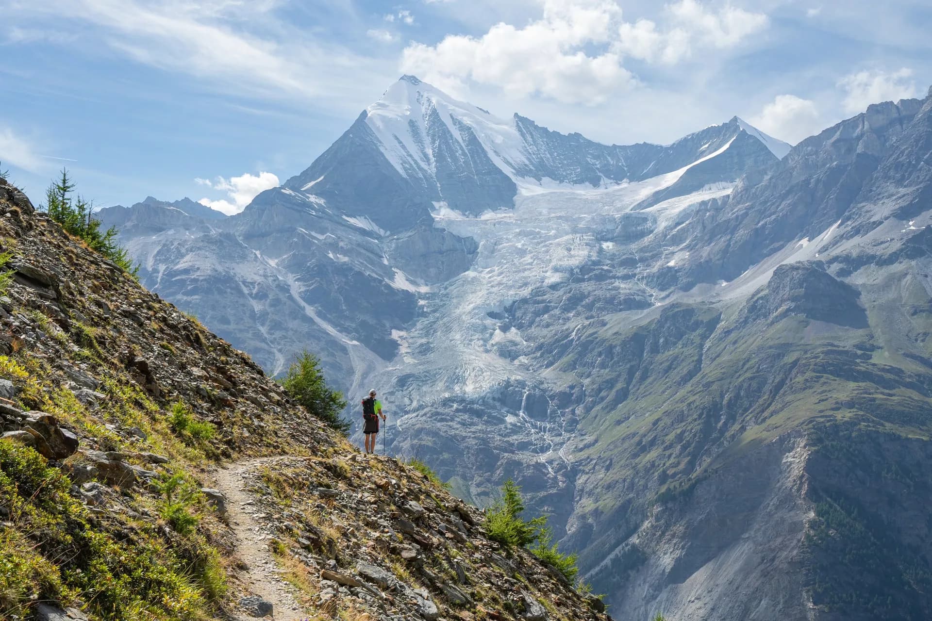 Hiker on rocky trail overlooking massive glacier and snow-capped Weisshorn mountain.