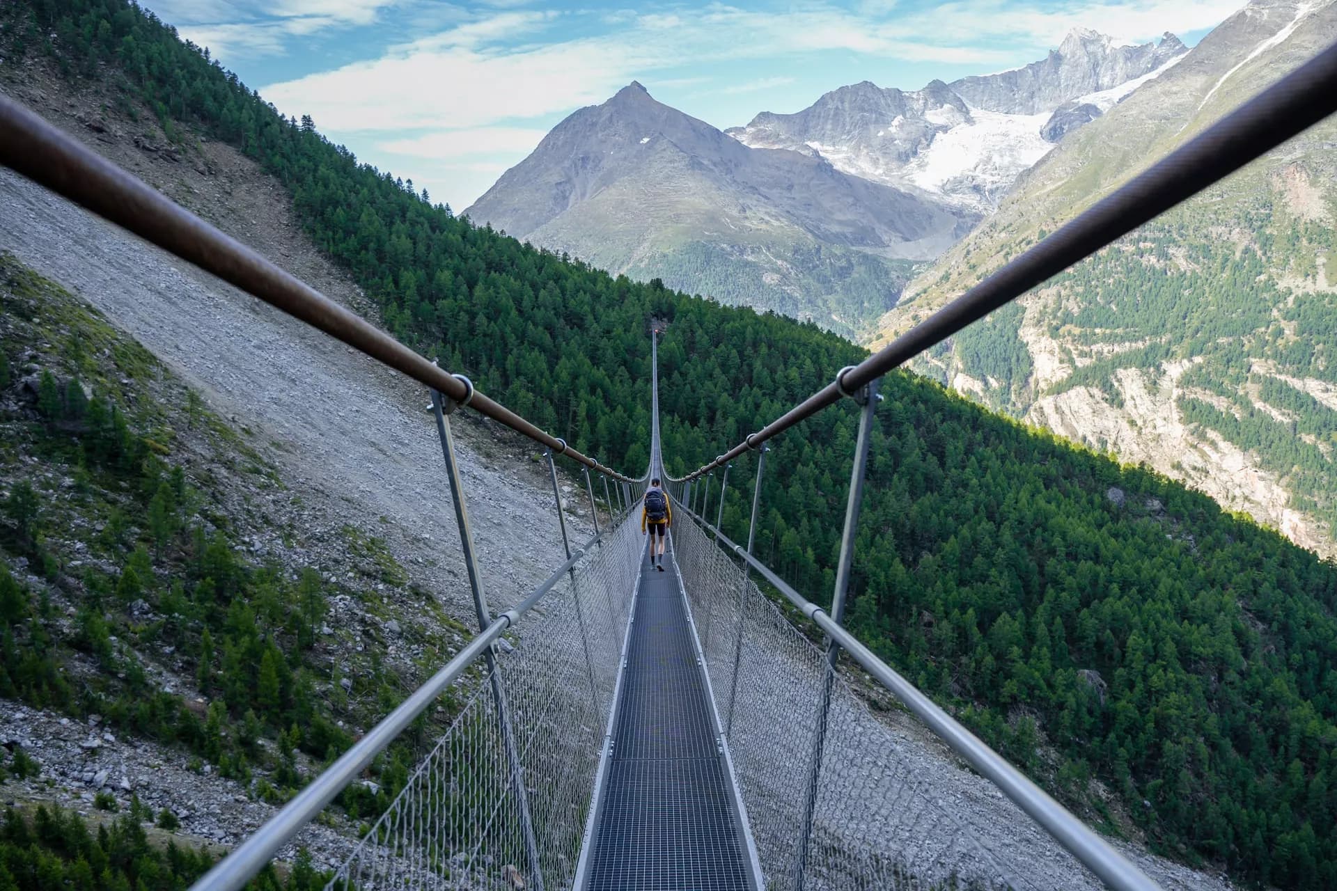 Hiker crossing the Charles Kuonen Suspension Bridge over a forested valley with snow-capped mountains.