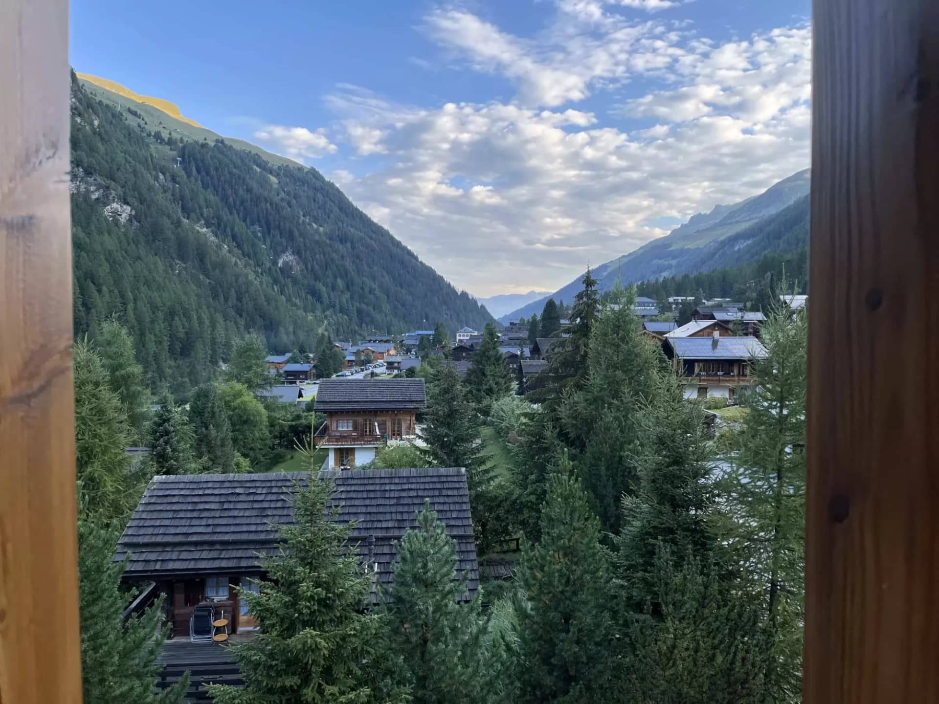 Alpine village nestled in a valley with wooden chalets and dense pine forests under a cloudy sky.