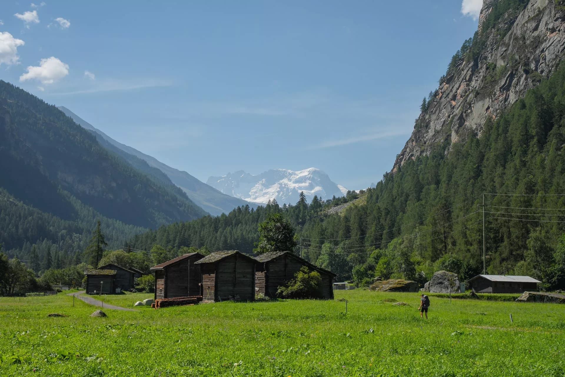Hiker walking past wooden huts in a green valley towards snow-capped mountains near Herbriggen.