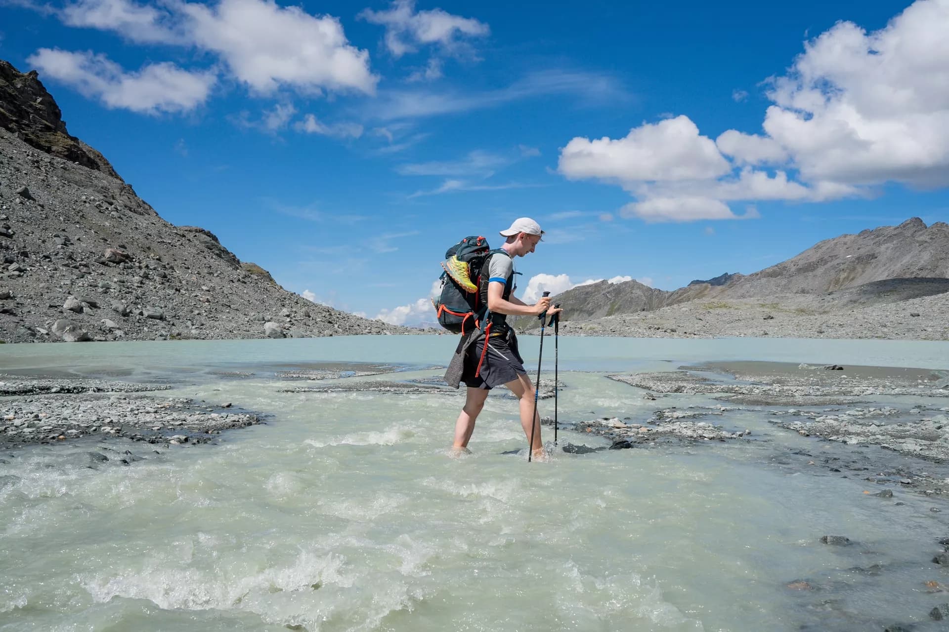 Hiker with backpack crossing rushing glacial stream near rocky mountains under blue sky.