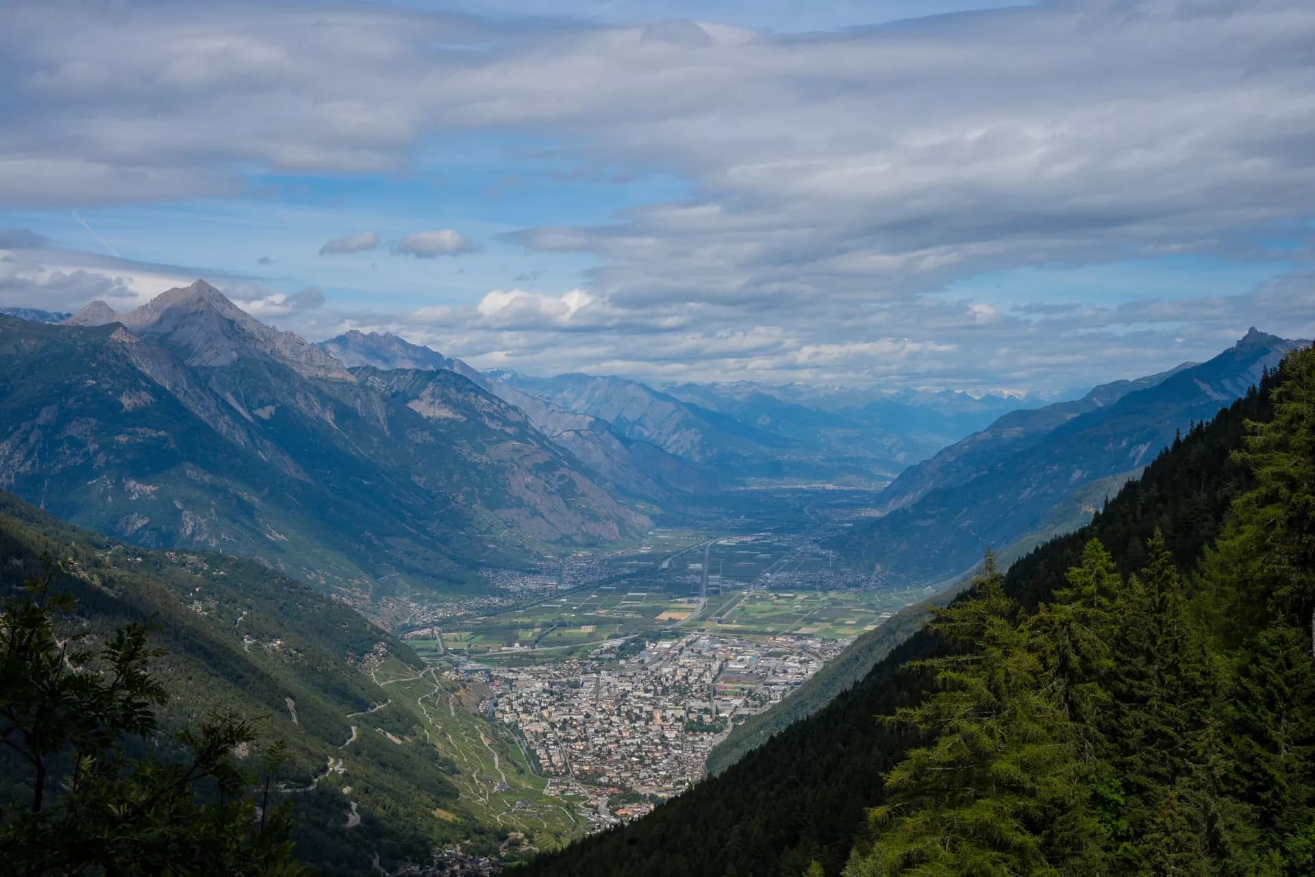 Panoramic view of a town in a valley surrounded by steep, forested mountains under a cloudy sky, Valais.