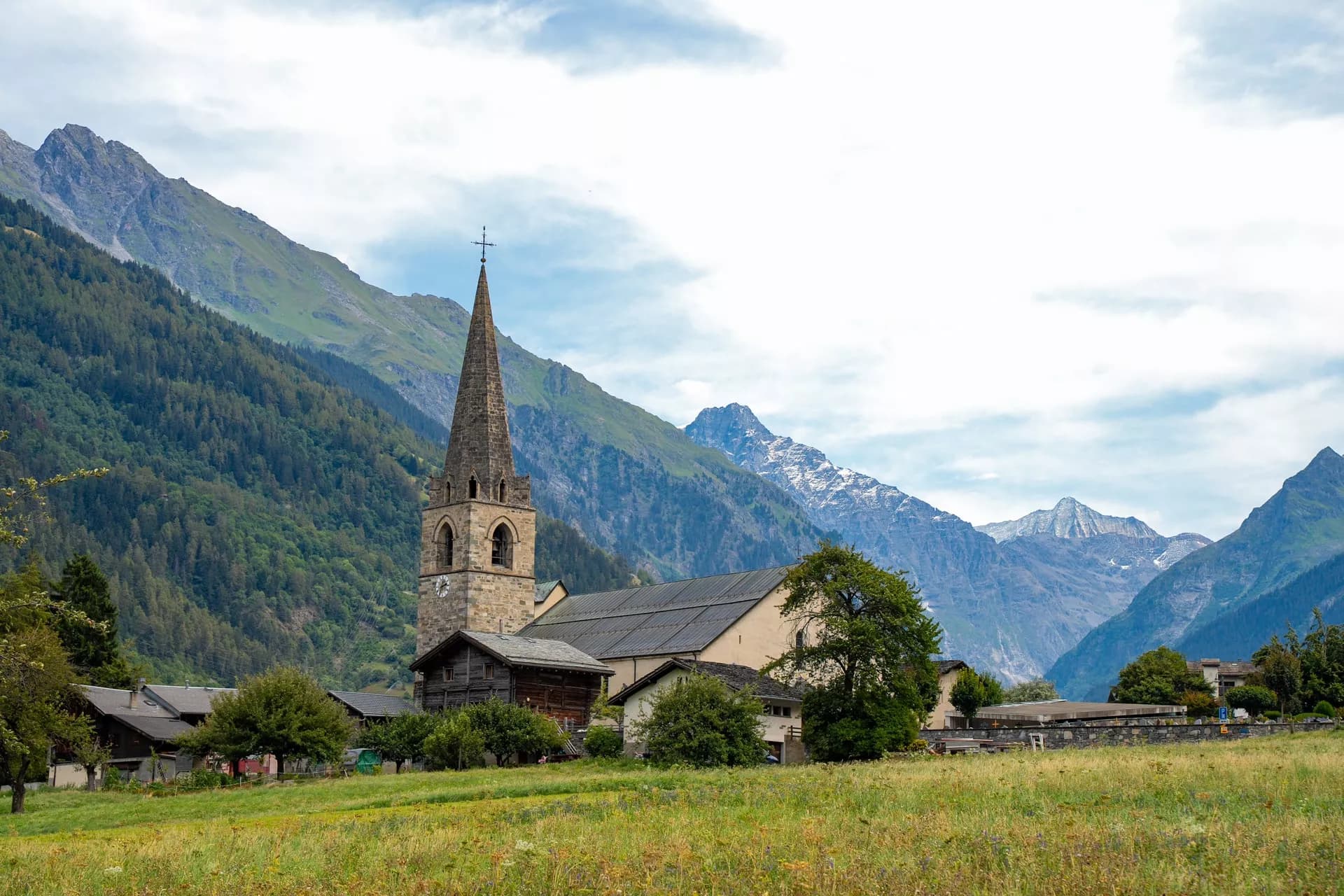 Stone church with tall spire in Le Châble village set against steep, forested mountains.