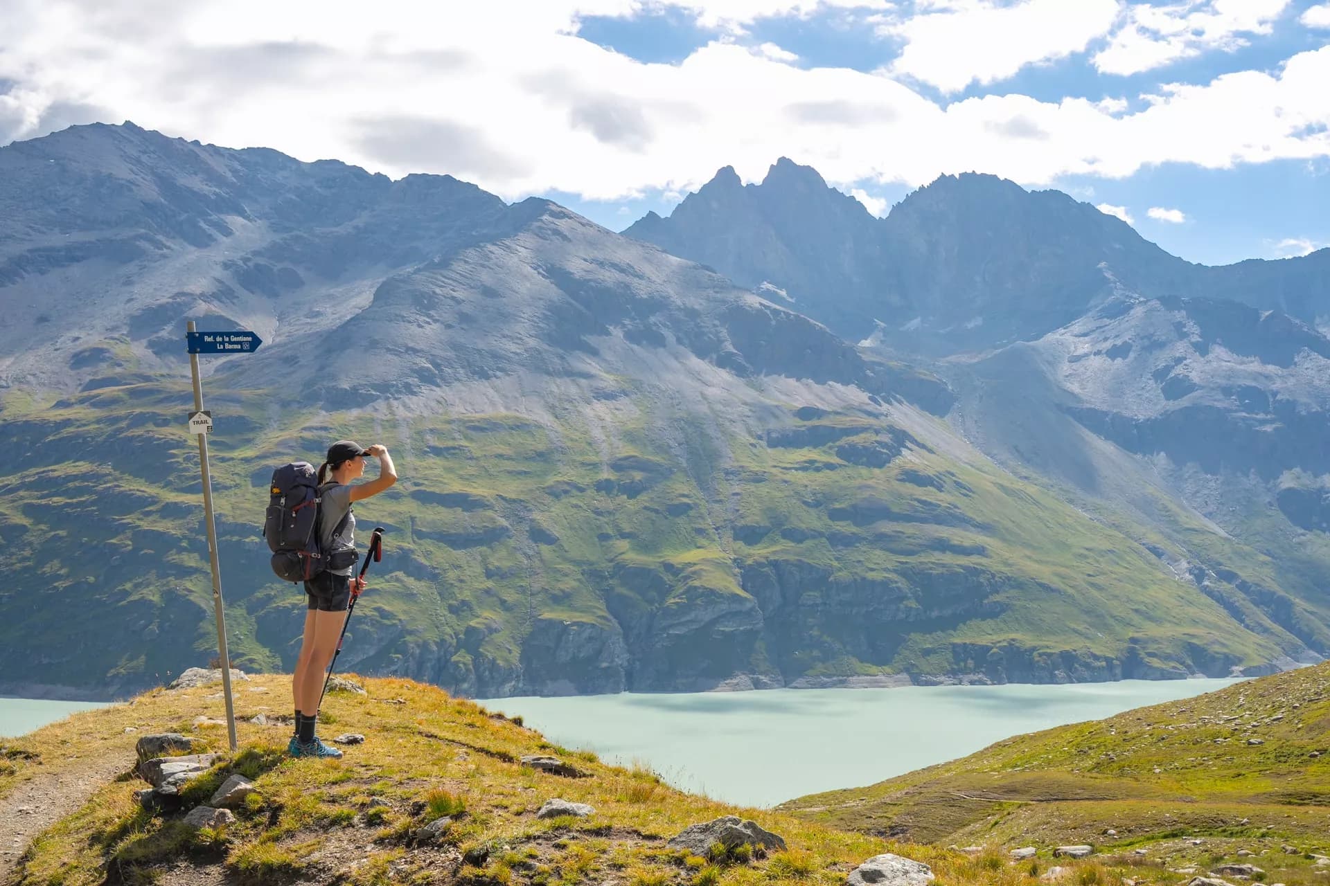 Hiker with backpack shading eyes overlooking turquoise Lac de Dix and steep mountains.