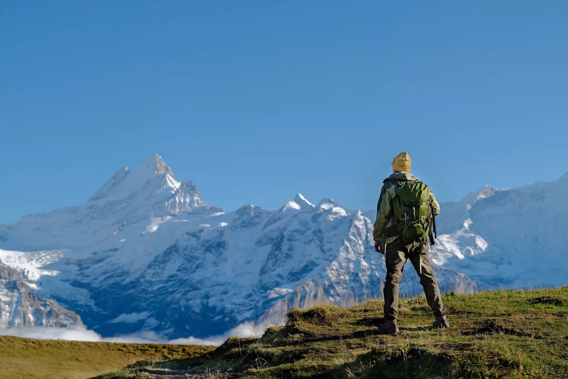 Hiker with backpack standing on grassy hill overlooking snow-capped mountains above Grindelwald.