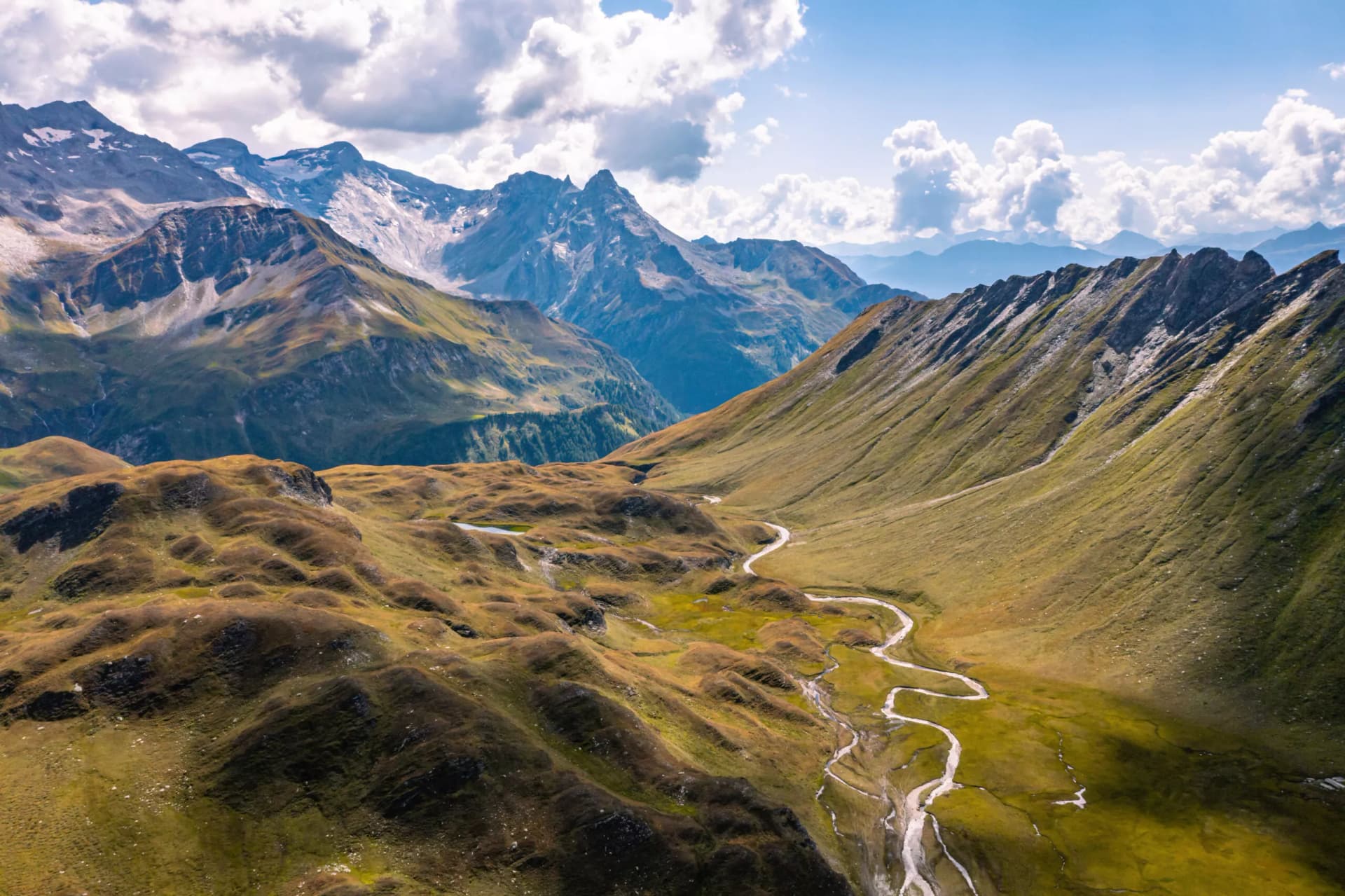 Aerial view of winding stream through grassy alpine valley toward snow-capped mountains under clouds.