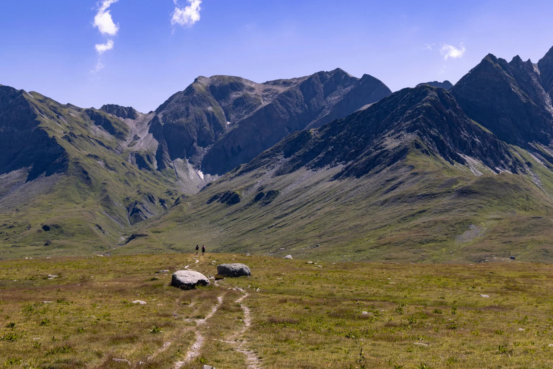 Two women walk on a winding alpine trail between mountains in late summer on a sunny day in the Greina Plateau, in Switzerland.