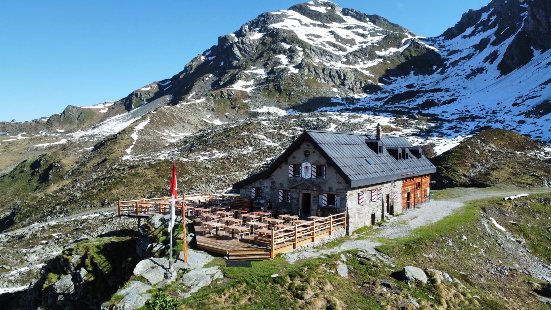 Stone mountain hut with outdoor terrace and Swiss flag in the Alps with snow-capped peaks.