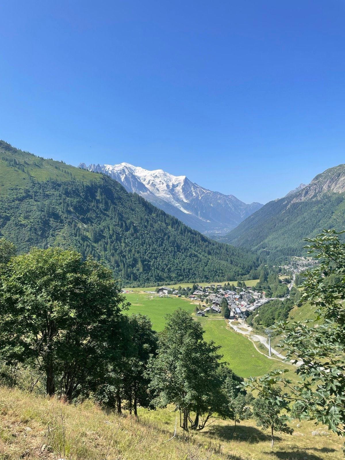 Alpine valley village with green slopes and snow-capped mountain peaks under blue sky