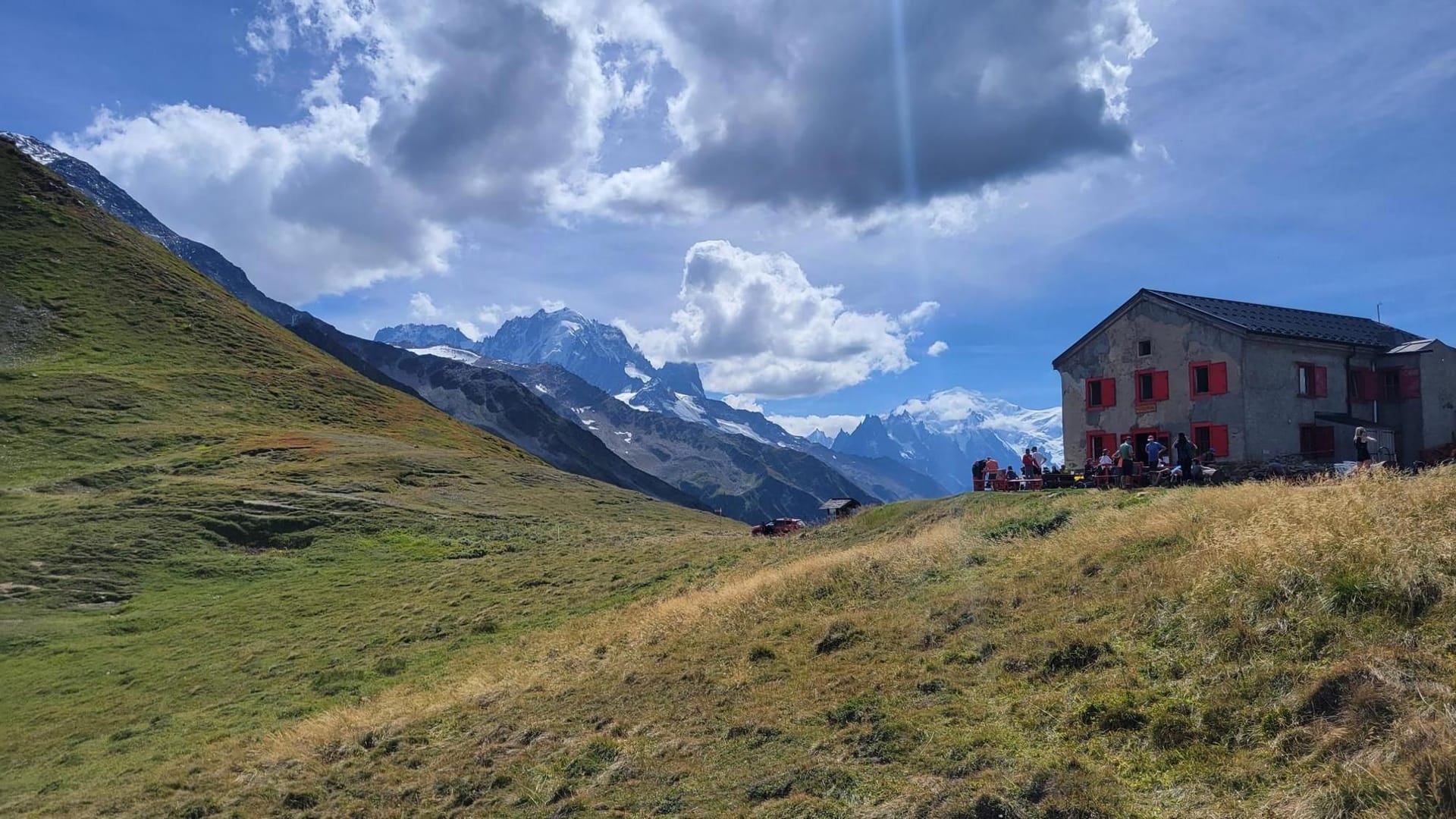 Alpine mountain refuge with people dining outside amidst snowy peaks and green slopes