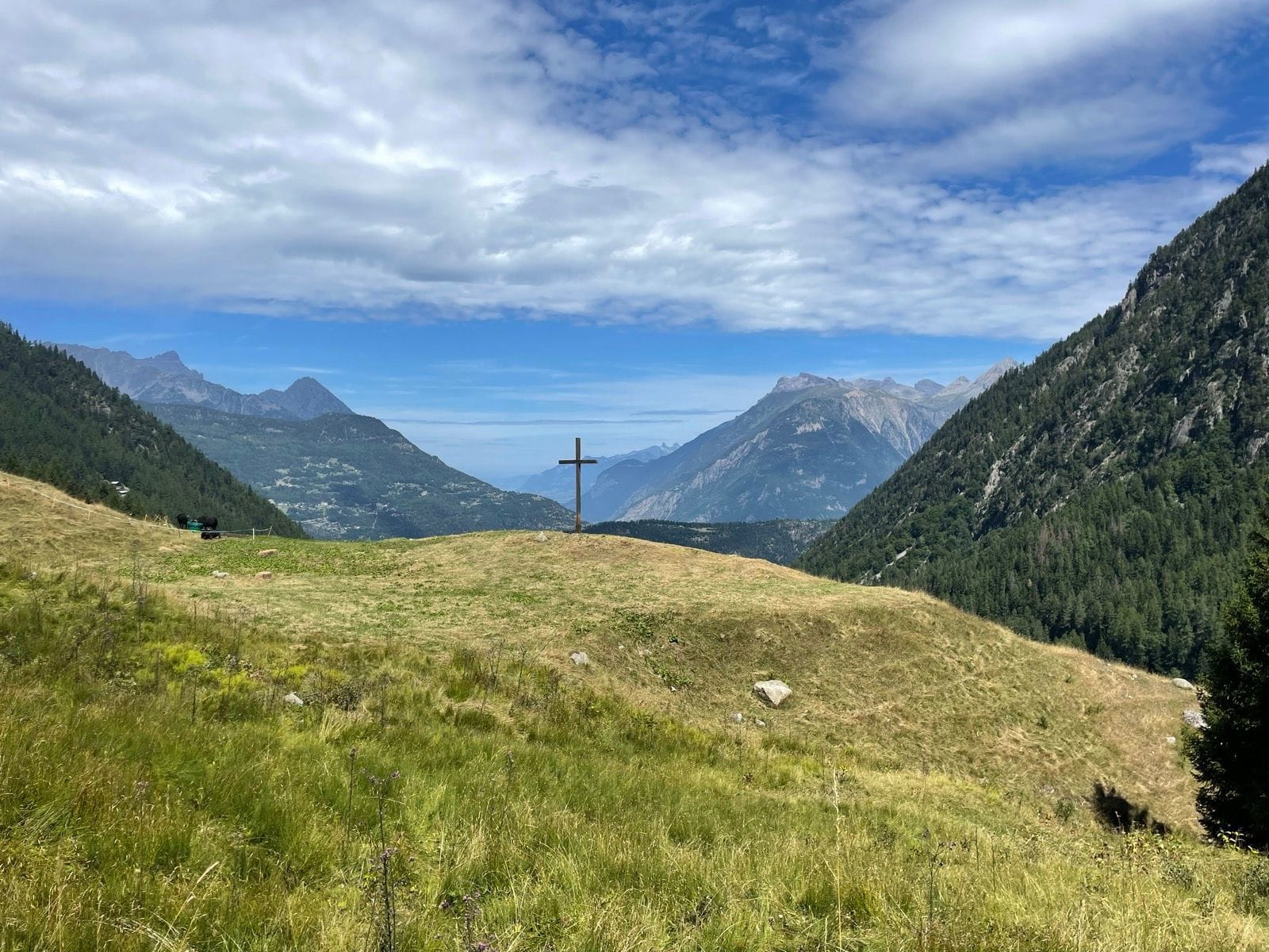 Wooden cross on grassy mountain ridge with alpine peaks and cloudy sky
