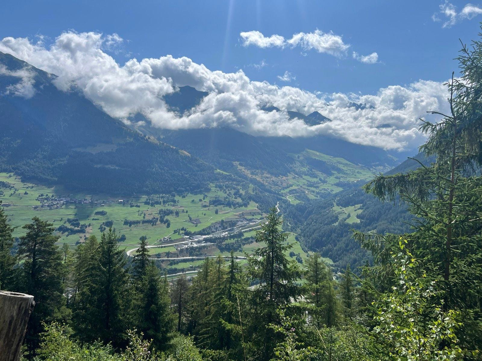Alpine mountain valley with green fields, winding roads, pine trees, and low-hanging clouds under a blue sky
