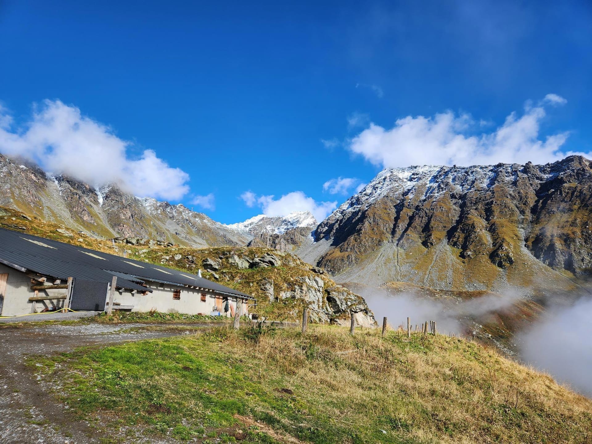 Alpine mountain landscape with a rustic building, snow-dusted peaks, and low clouds under a blue sky