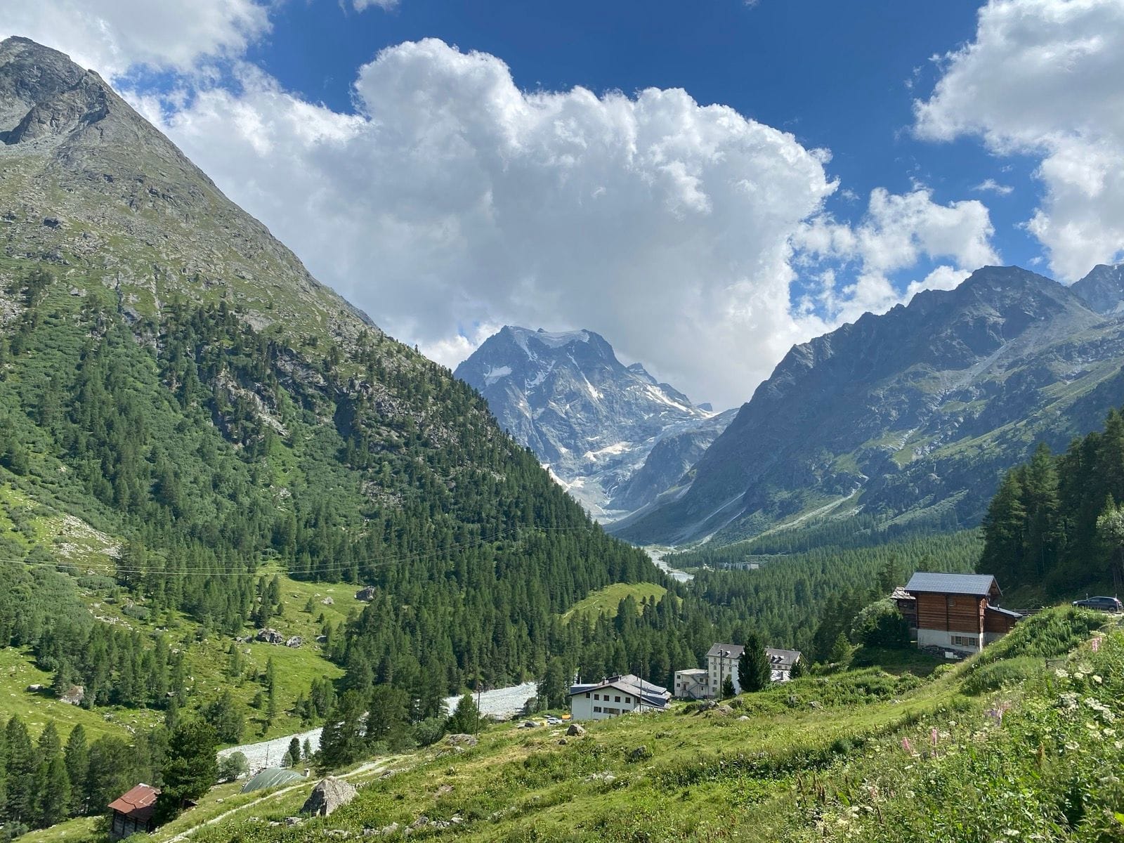 Alpine mountain valley with pine forests, river, and buildings under cloudy sky