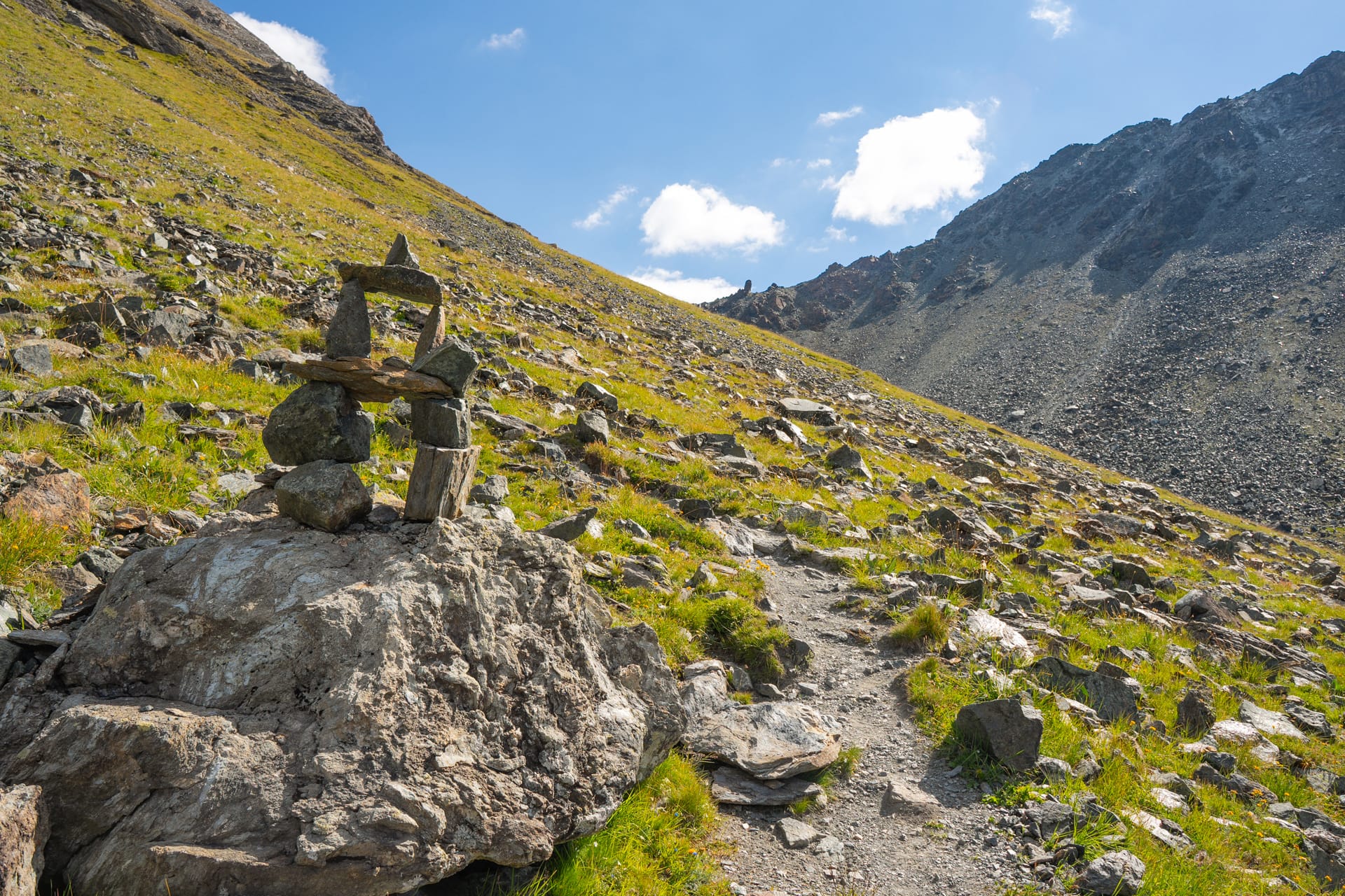 Stone cairn beside mountain hiking trail on grassy slope under blue sky