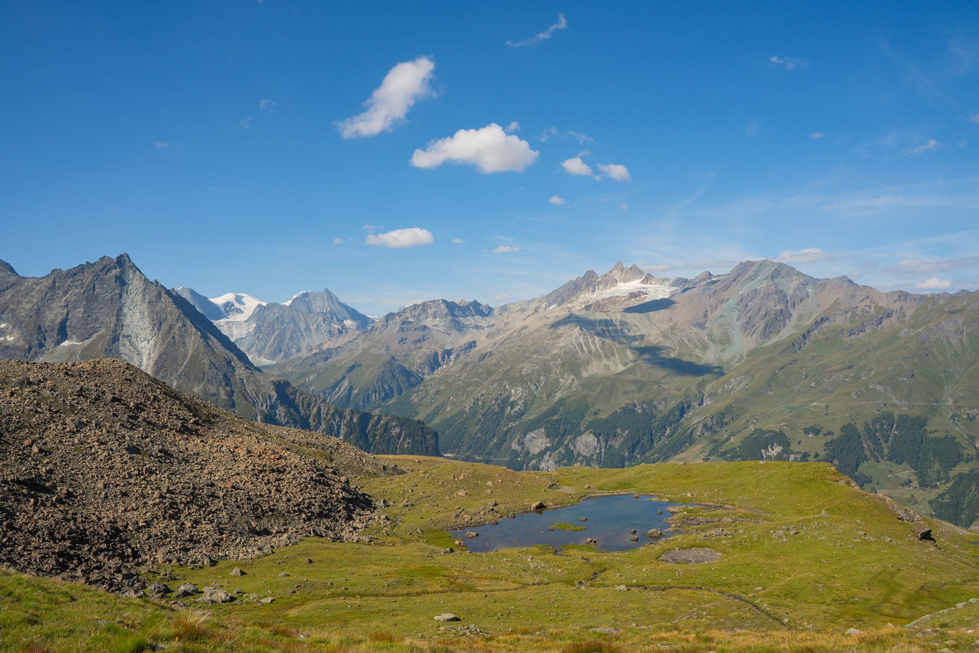 Alpine mountain landscape with rocky peaks, green slopes, and small tarn under a blue sky with white clouds