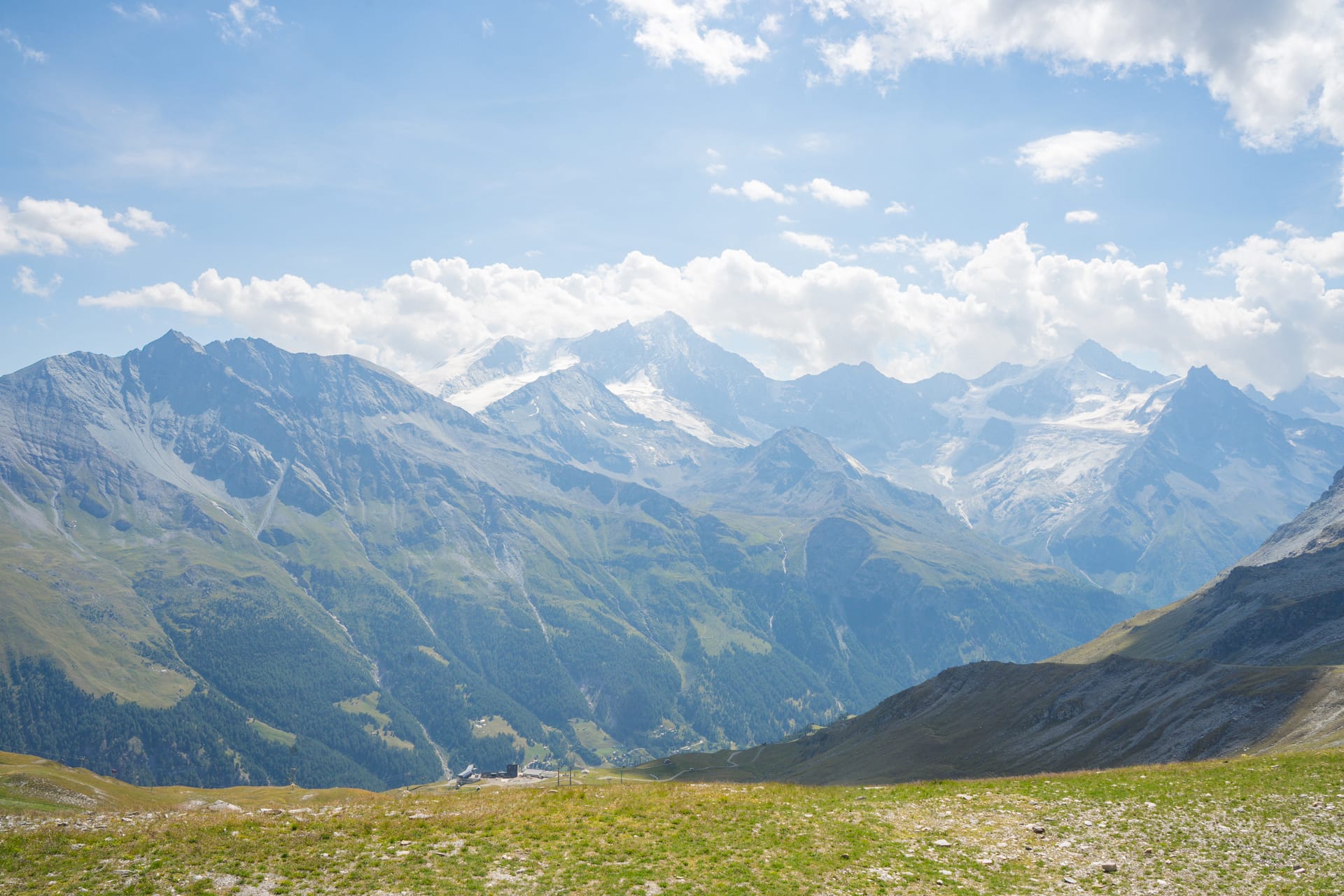 Alpine mountain range with snow-capped peaks and grassy foreground under a cloudy sky