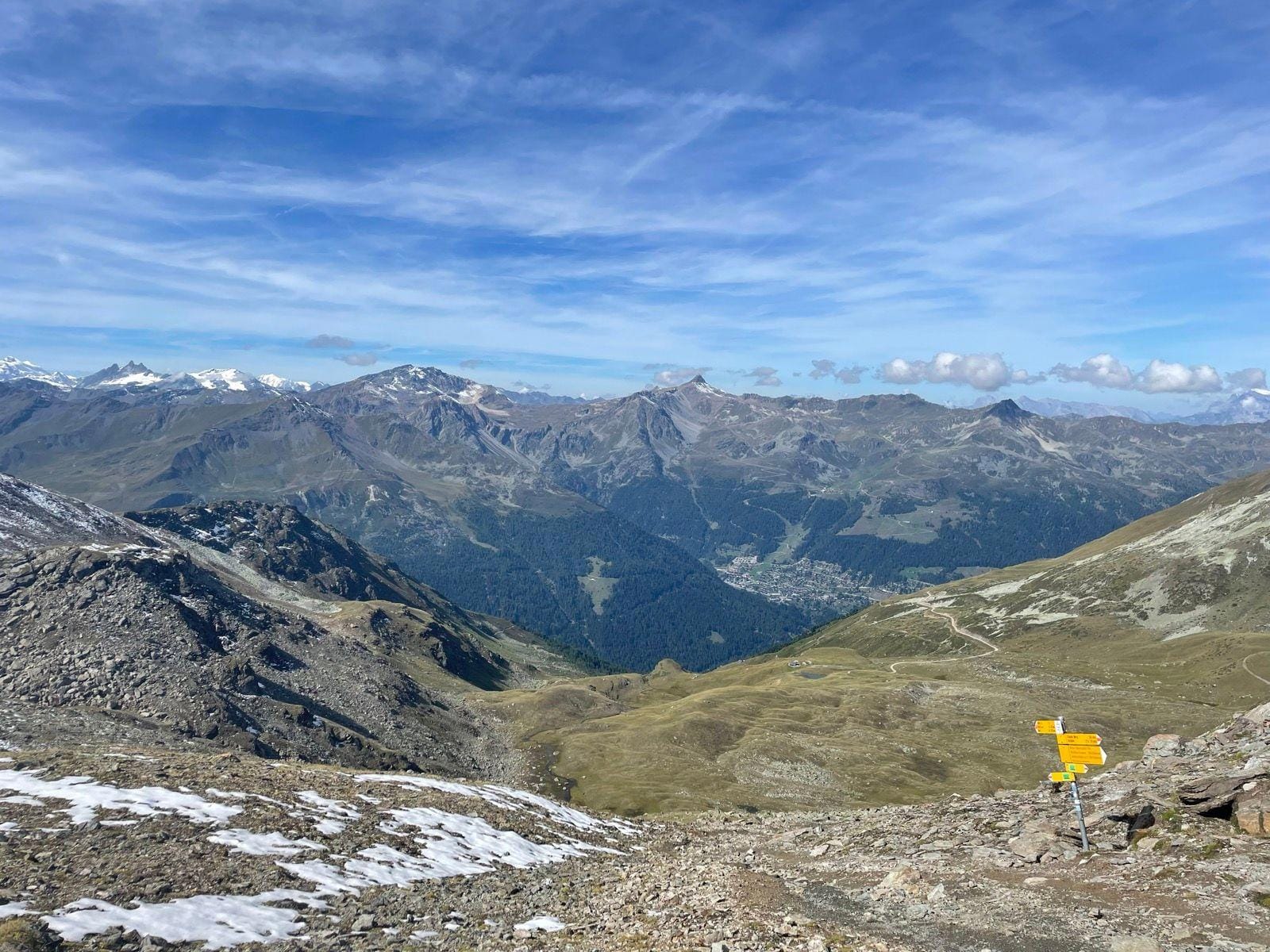 Alpine mountain landscape with hiking trail signs, rocky slopes, and distant valley under a blue sky