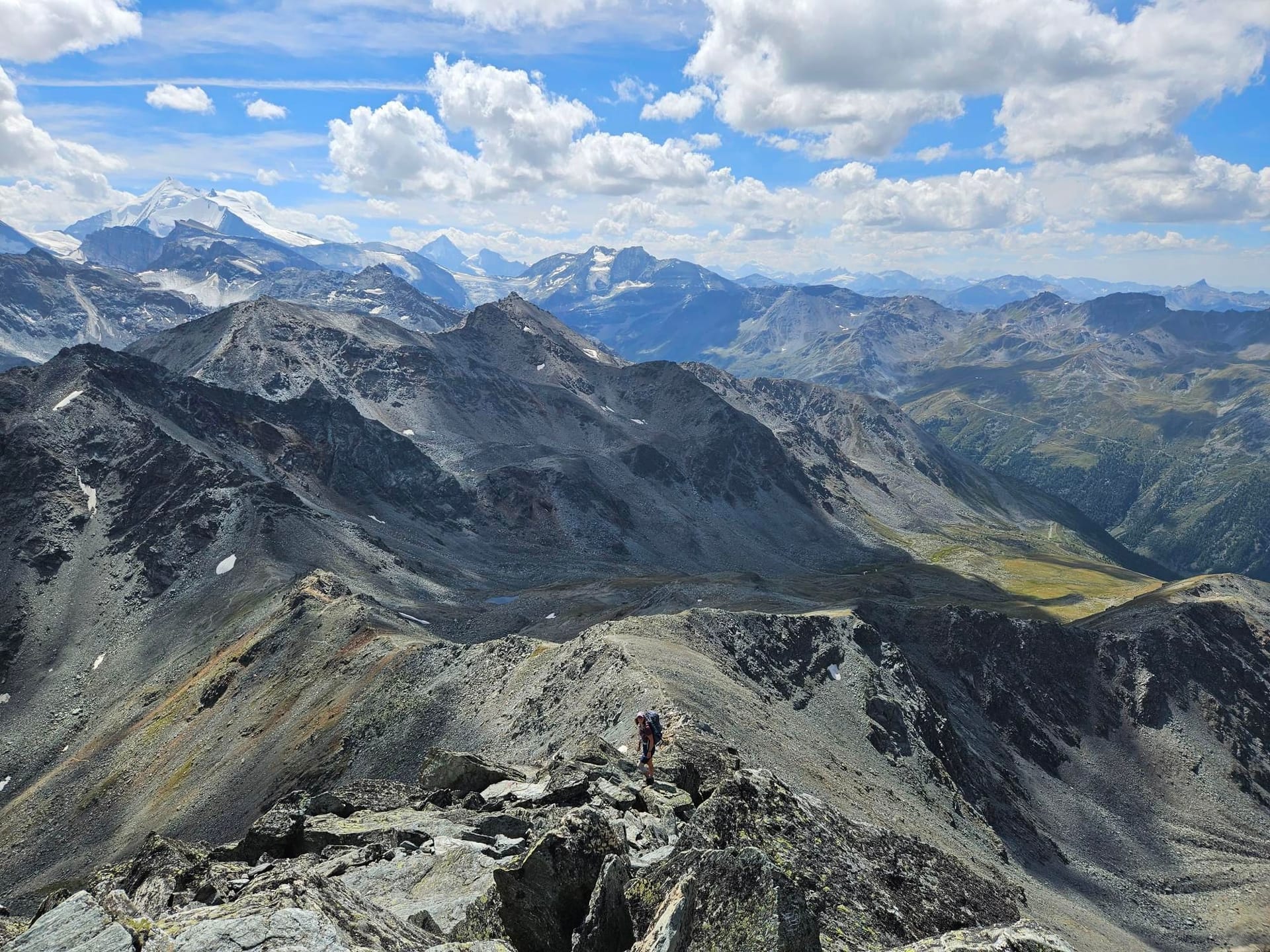 Hiker walking along a rocky mountain ridge with panoramic alpine views and cloudy blue sky