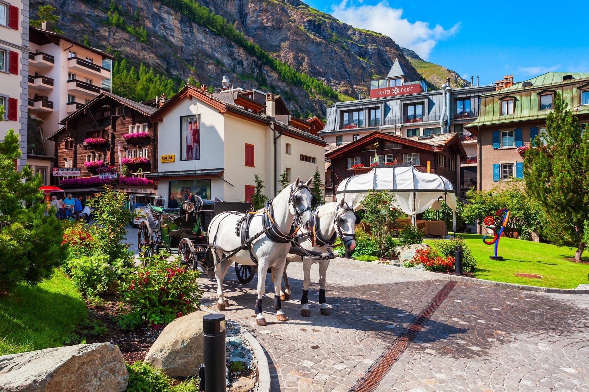 White horses pulling a carriage on a cobblestone street in Zermatt with alpine buildings and mountains