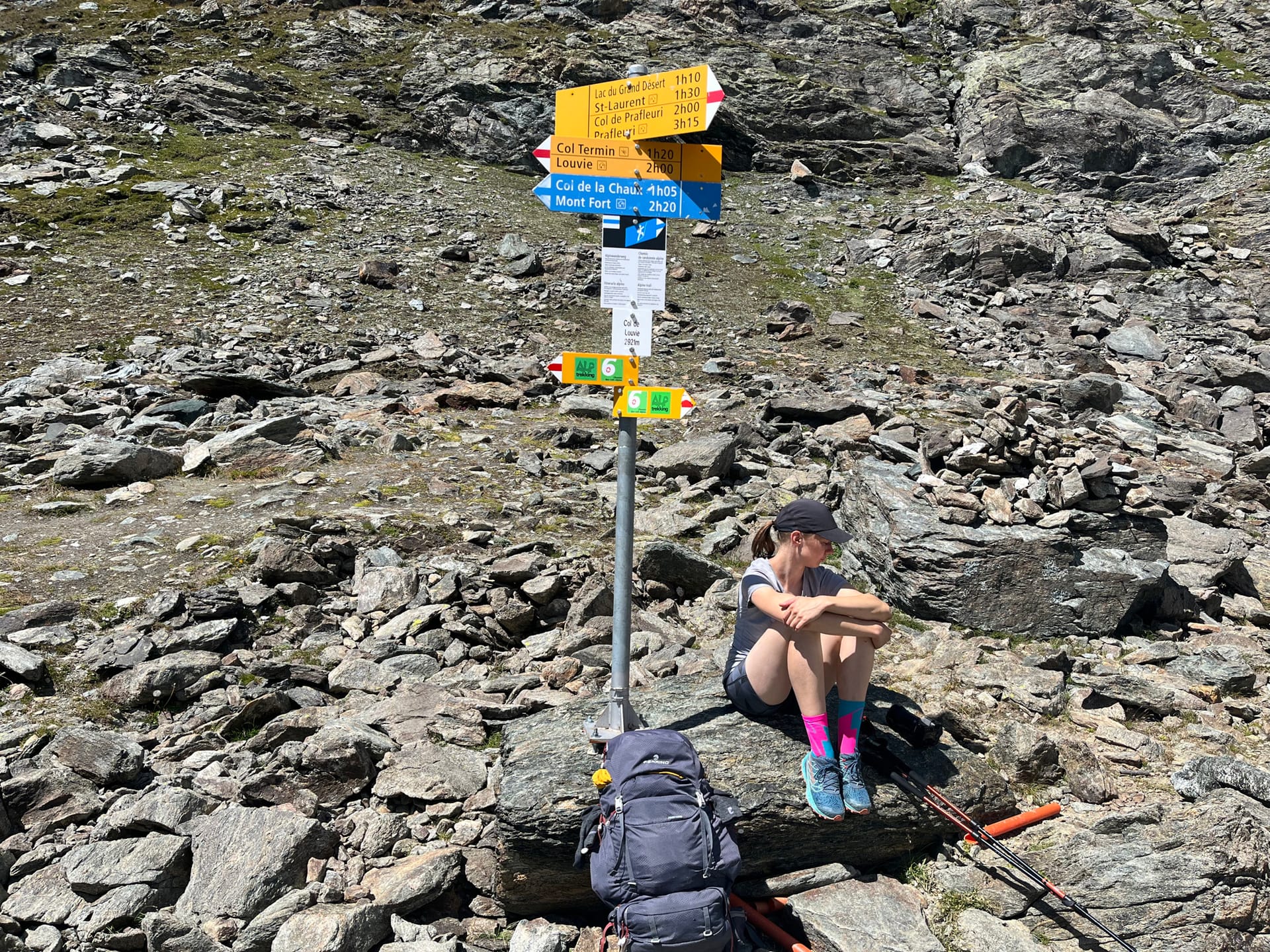 Hiker sitting on rock next to hiking trail signpost in rocky alpine terrain