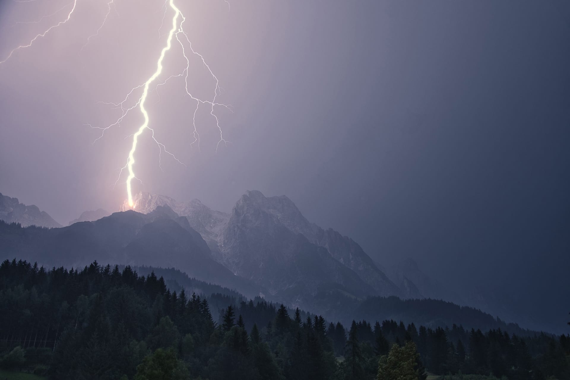 Lightning bolt striking mountain peaks over pine forest during storm