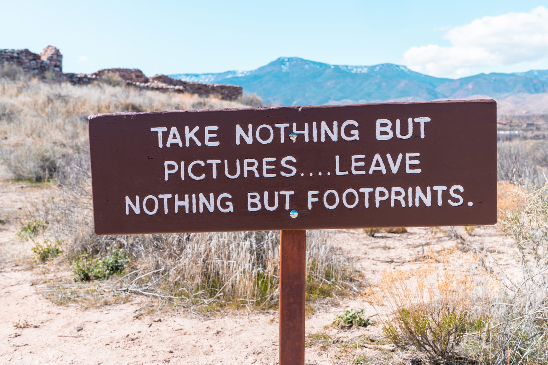 Brown wooden sign with white text reading take nothing but pictures leave nothing but footprints outdoors in desert landsc...